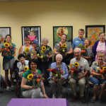Photo of a group of memory café attendees holding up their flower arrangements and all smiling at the camera.