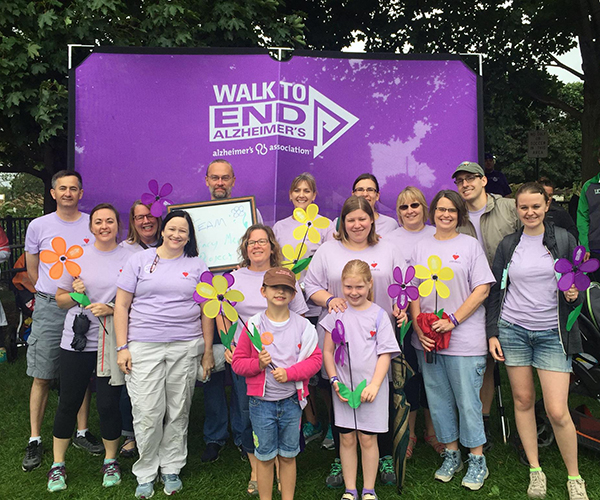 A group of people in matching purple shirts standing together and all smiling at the camera in front of a sign that reads Walk to End Alzheimer's