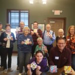 Photo of a group of memory café attendees holding up their painting crafts and all smiling at the camera.
