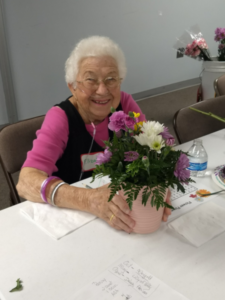 An elderly woman sitting at a table holding a pot of beautiful flowers