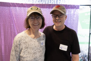 An older couple wearing tan baseball caps pose in front of a sparkling, purple curtain.