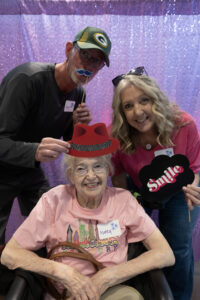 An older woman in a wheel chair smiles as a man and a woman hold up silly props for the camera.