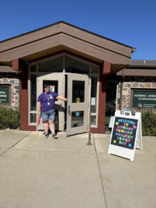 A person in a purple shirt poses at the front of a building. An A-frame out front says "Welcome to Library Memory Project Family Day."
