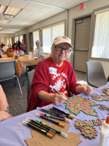 A woman in a red shirt and cardigan decorate a cardboard maple leaf.