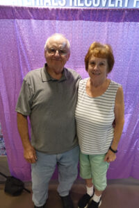 An older couple poses in front of a purple party screen.