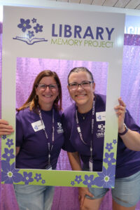 To library workers in purple Library shirts pose inside a frame for the Library Memory Project.