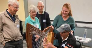 A group of older adults surround a gentleman playing a harp.