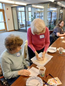A woman with the nametag Carol demonstrates a craft in a white container to a woman with the nametag Phil