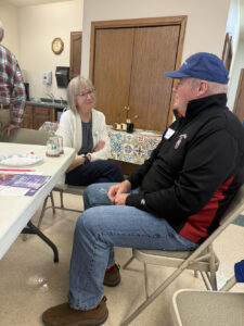 A man in a black jacket speaks with a woman in a white cardigan. Both are sitting at a table corner.