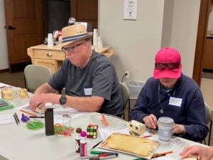 A man and woman wearing hats decorate a small treasure box.