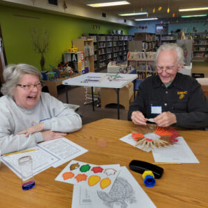 A man in a black shirt makes a clothespin wreath while the woman next to him laughs.