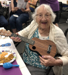 An older woman in a white cardigan smiles as she holds a ukelele