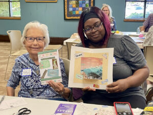 An older woman and a younger woman show off their collage craft that they made at a Memory Cafe.