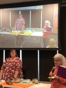 An older woman with curly white hair speaks into a microphone next to a woman about to do a culinary demonstration.
