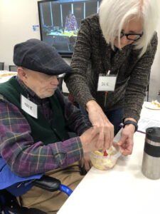 A woman with the name tag Sue assists a man with the name tag Ken E. with stirring something in a plastic container.