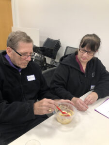 A woman in a black sweatshirt assists an older man with stirring something in a round, plastic container