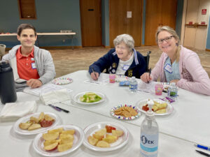 An older woman with a woman and man on either side of her sit at a table with plates of sliced apples and one plate of cookies.