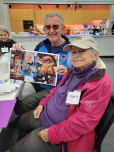 A woman in a pink jacket smiles for the camera while a man in sunglasses holds up a collage of animals.