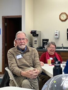A gentleman in a brown sweater listens to a presenter at an event.