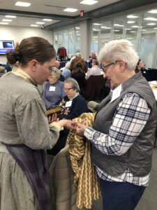 A woman in a period costume talks about a blanket held by a woman in a plaid shirt and vest.
