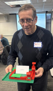 An older gentleman in a blue sweater with "Bill :)" on his name tag holds a lego construction.