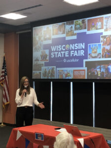 A woman talks at a presentation. Behind her is a projection for the Wisconsin State Fair.