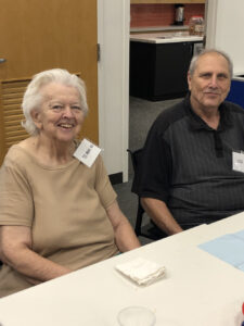An older woman in a beige shirt and a man in a dark polo smile for the camera.