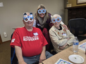 An elderly couple and a younger woman behind them smile for the camera as they wear fun snow day themed glasses.