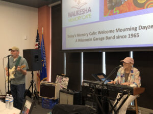 Two performers, a man at a guitar and a woman at a keyboard, play in front of a projection that reads "Today's Memory Cafe: Welcome Mourning Dayze, A Wisconsin Garage Band since 1965"