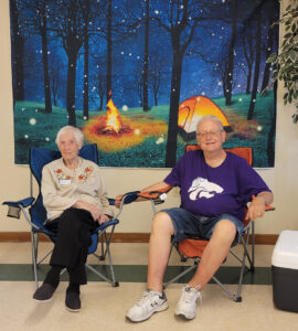 An older couple sit in lawn chairs in front of a camping backdrop.