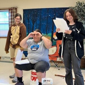 A woman sitting on a bucket mimes binoculars with her hands while two young women smile on either side of her.