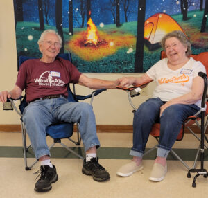 An older couple sit in lawn chairs and hold hands. Behind them is a backdrop of a tent and campfire in a forest.