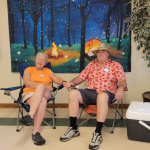 An older couple sit in lawn chairs in front of a backdrop of a tent and campfire in a forest