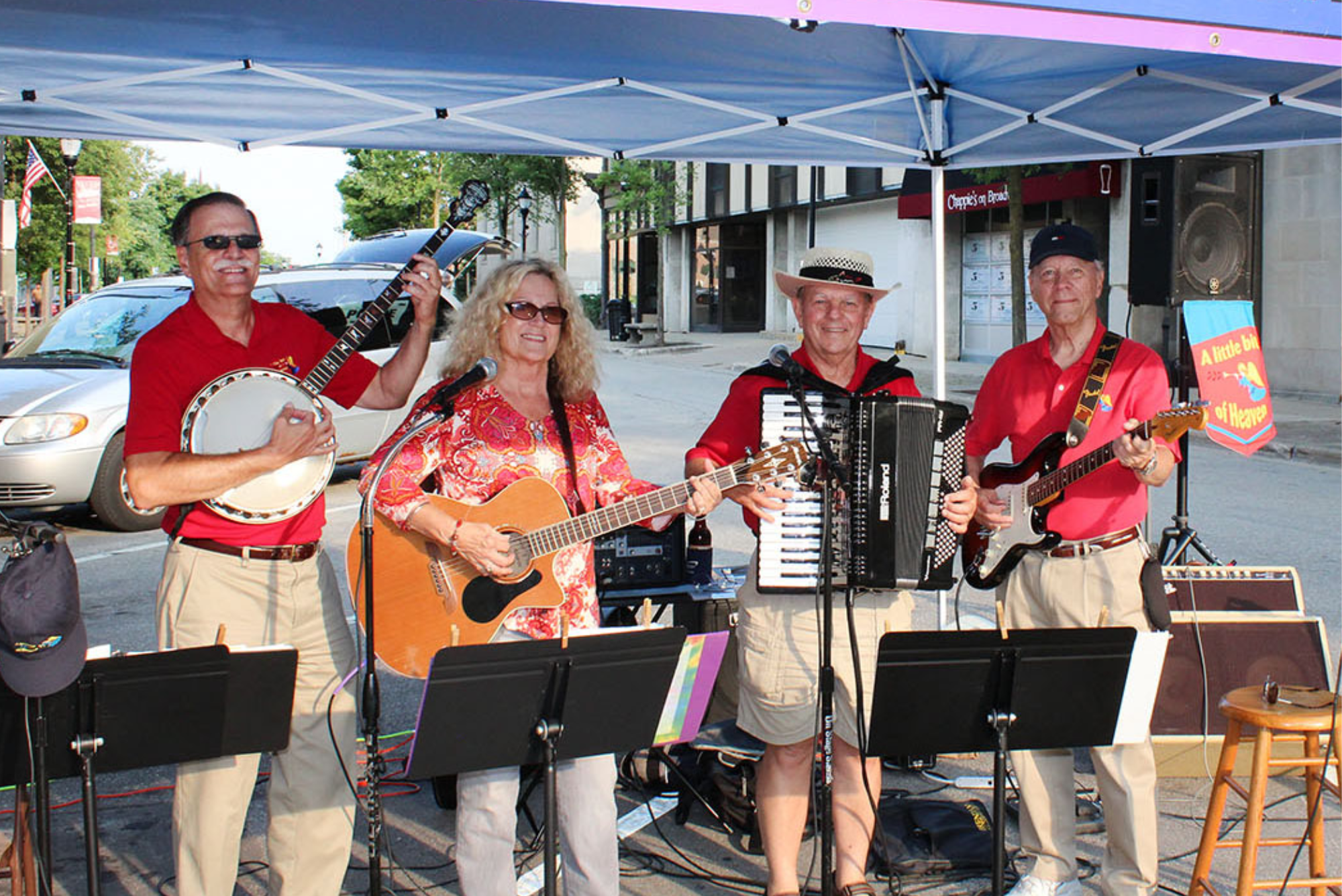 A four person band plays at an outdoor festival. From left, there's a man with a banjo, a woman with a guitar, a man with an accordion, and a man with an electric guitar.