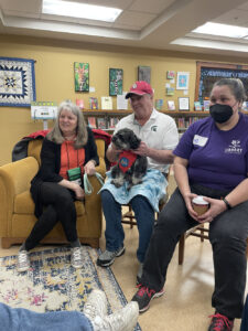 Three people gather around someone off-camera during an event. The man in the middle pets a dog on his lap.