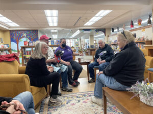 A group of five people gather around a lounge area at a library. A man wearing a red cap pets a dog on his lap. One of the other people is a Library Memory Project worker. The other three wear black and are engaged in a conversation.
