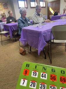 A group of people play bingo while sitting at tables with purple tablecloths