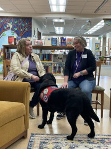 Two women have a conversation in a library while a black service dog in a red vest stands at their knees.