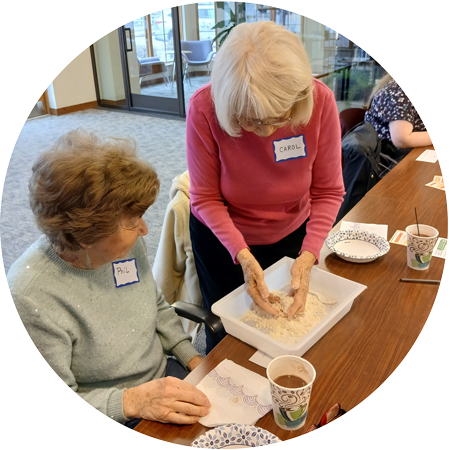 An older woman sifts sand or flour with her hands while a sitting woman watches