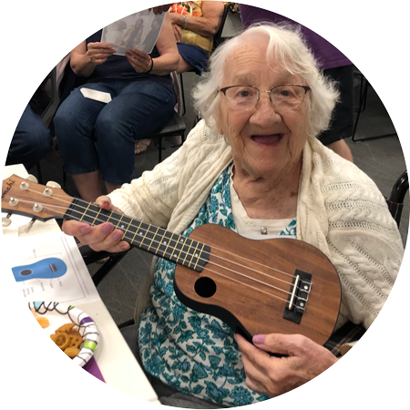 An elderly woman sitting and playing a ukulele with a large smile