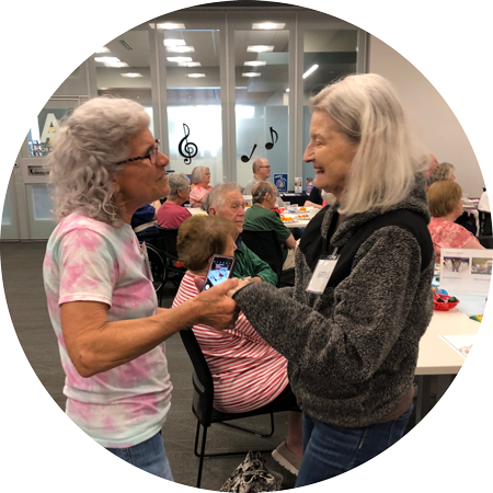 Two older women standing facing each other and holding both hands, there are people sitting at tables behind them