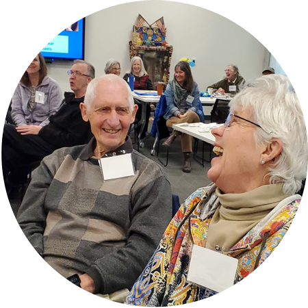 An elderly woman and man looking at each other and laughing, there are people sitting at tables and smiling behind them