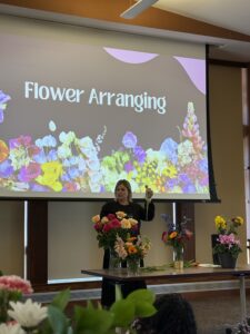 Woman demonstrating flower arranging with various flowers in vases on a table.