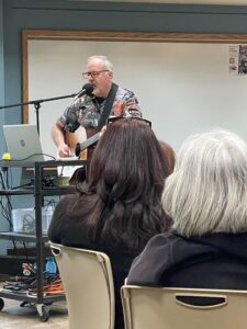 A man plays guitar and sings into a microphone in front of a whiteboard with seated audience members watching.