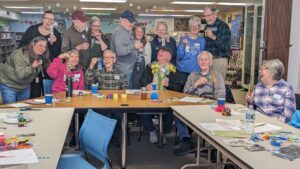 A group of older adults in a library smiling and showing off handmade bookmarks at a craft table.