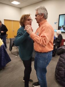 Two older adults are dancing closely in a room with others seated nearby.