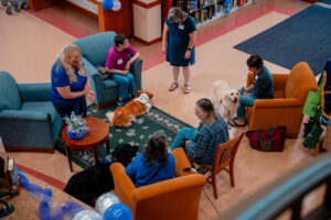 Three dog handlers and their dogs sit in chairs and visit with others