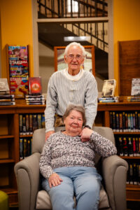 Husband stands behind wife who is sitting in a chair as they hold each others hands