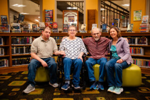 A family of four adults sit together with library books behind them.