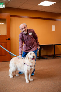 An older man with glasses and a dog indoors, standing on a carpet.